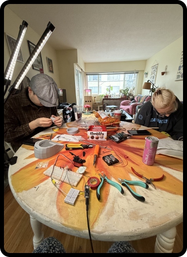 Both team members working at the build table — tools, soldering iron, breadboard, electronics